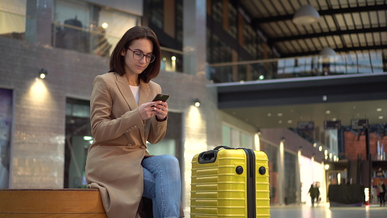 A young woman sits with a phone in her hands in the airport with a suitcase. Girl in pink coat. 4k