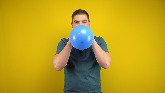 A young man inflates a blue balloon with his mouth on a yellow background. Man in a green polo. 4k