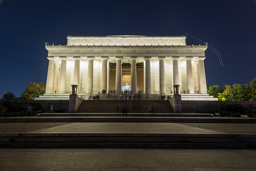 Tourists visit the Lincoln Memorial in Washington, D.C. on an autumn night. Long exposure wide shot from the east side of the building. The lights of a passing airplane are seen in the sky.