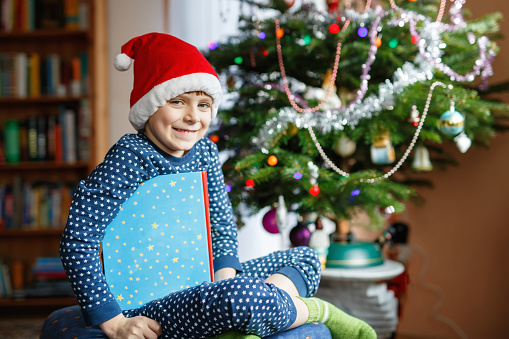 Beautiful little school kid boy reading a book near Christmas tree with lights and illumination. Happy child with Santa Claus hat having fun with book gift. Celebrating family holiday. Education
