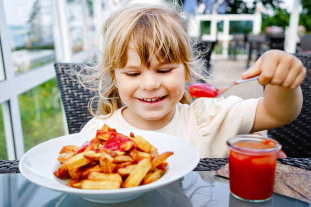 Portret szczęśliwej uśmiechniętej przedszkolaczki jedzącej frytki z ketchupem pomidorowym w restauracji na tarasie na świeżym powietrzu. Małe dziecko z blond włosami lubi niezdrowe fast foody lub świeżo przygotowany lunch. – zdjęcie