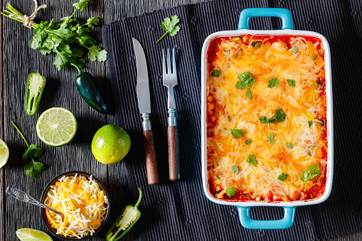 vegetarian enchilada casserole layered with tortillas, sauce, pinto beans, corn, zucchini and mexican blend cheese  in baking dish on dark wood table, landscape view from above, flat lay