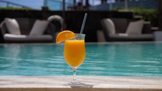A close-up of a glass of freshly squeezed orange juice with an orange slice on the side of the glass and a metal straw, standing on the very edge of the pool.