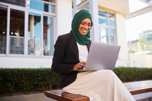 Young muslim businesswoman wearing a hijab smiling while sitting on a bench outside of her office building and working on a laptop