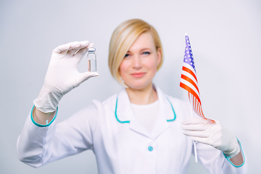 A female doctor wearing protective gloves holds a vaccine in a test tube that was won from America, ready to be tested for efficacy against the coronavirus. American doctor with usa flag before vaccination healthcare, medicine and pharmacy concept