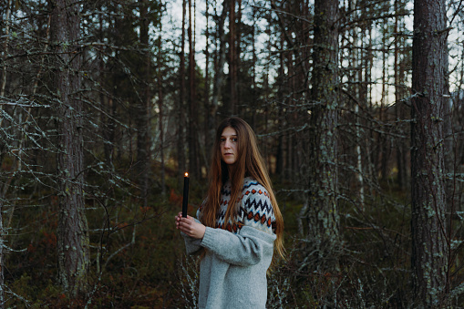 Side view of female with long hair in knitted sweater walking in the spooky woodland during the dark evening time holding candle in Norway