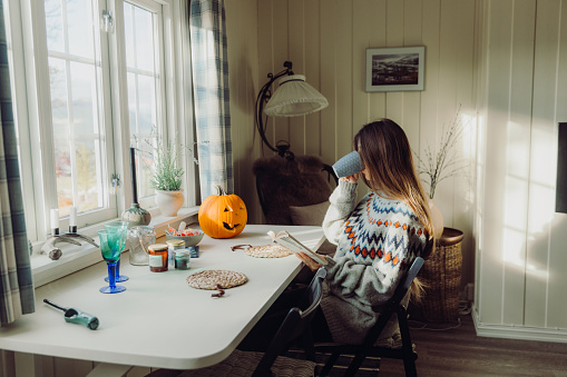 Female with long hair in knitted sweater sitting at the table with mountain view, reading book and drinking coffee enjoying the rural autumn vibes in the hut