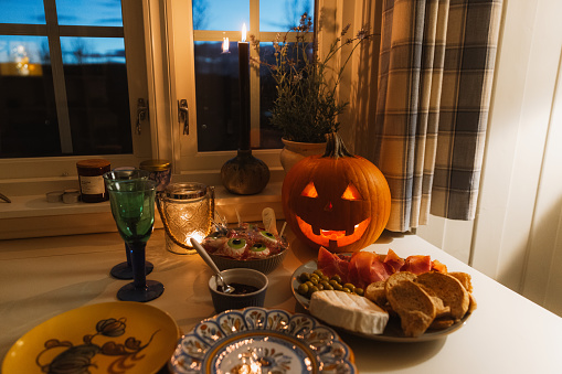Celebrating halloween with a dinner with cheese and meet appetiser and candies served on the colorful plates, with lighting Jack O'Lantern and window view of the mountains in Norway