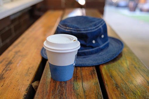 A takeaway coffee and a blue denim hat on a wooden bench.