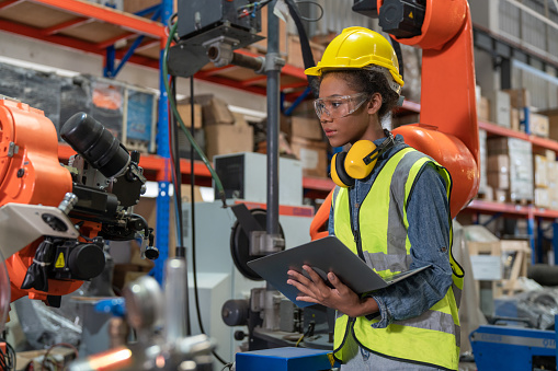 Young African American female engineer with tablet operating welding robot arm machine in warehouse factory.Professional technician automation robotic industrial