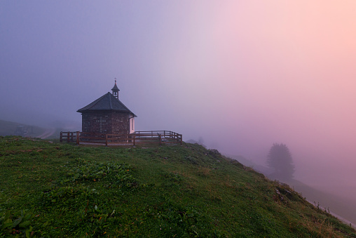 The reddish light of the sunset colors the thick foggy clouds at the chapel of the Ackernalm, Tyrol, Austria