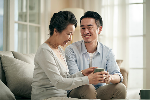 asian adult son sitting on couch at home chatting with senior mother happy and smiling