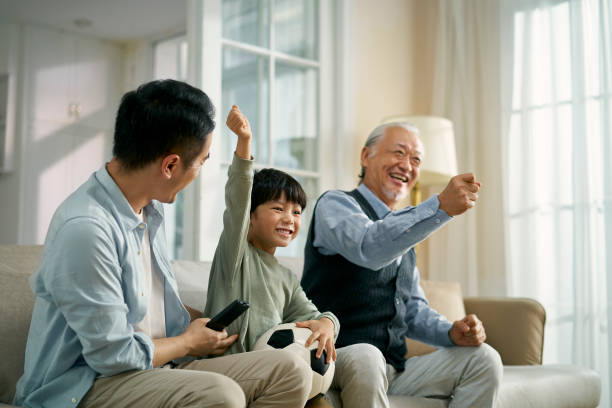 asian son father grandfather watching soccer game on TV together at home asian son father grandfather sitting on couch at home celebrating goal and victory while watching live broadcasting of football match on TV together asian kids watching tv stock pictures, royalty-free photos & images