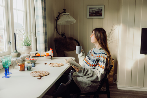Female with long hair in knitted Scandinavian sweater enjoying the sunny morning sitting by the table with window view and reading book with a cup of coffee, Norway