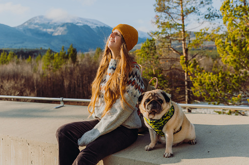 Smiling female with backppack, in yellow hat sitting at the viewpoint with a beautiful pug, admiring a view of the snowcapped mountain peaks and shiny autumn forest in Highland Norway