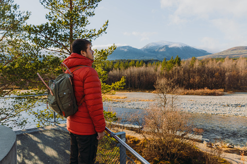 Side view of a male backpacker in red jacket staying at the viewpoint admiring a view of the snowcapped mountain peaks, shiny autumn forest and the river in Highland Norway