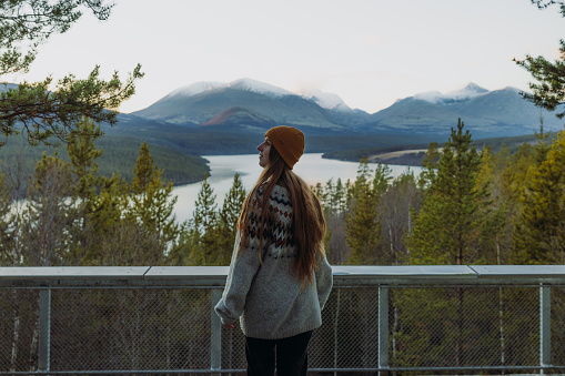 Side view of female with long hair in yellow hat staying at the viewpoint admiring the scenic mountain landscape with lake and the pine forest during sunset in Rondane National Park