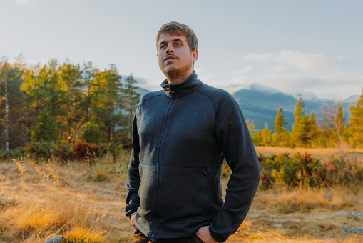 Hipster traveler in a down jacket and sunglasses stands on a mountain slope against the backdrop of epic rocks and smiles. The concept of finding happiness in the mountains.