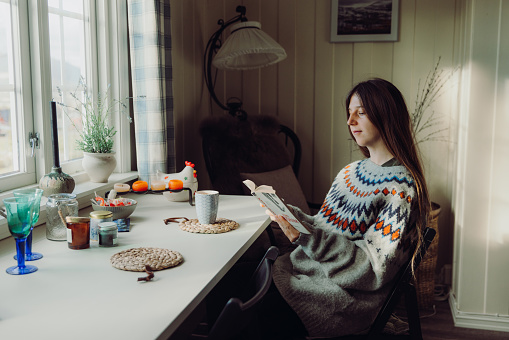 Female with long hair in knitted Scandinavian sweater enjoying the sunny morning sitting by the table with window view and reading book with a cup of coffee, Norway