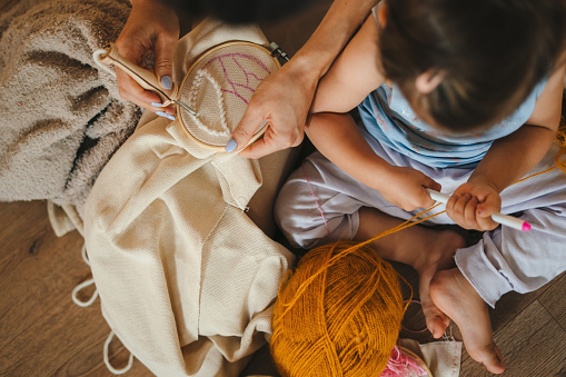 Top view of a mother and little baby daughter sitting on the floor in living room knitting with needles together. Family activity at home. People lifestyle. Happy family.