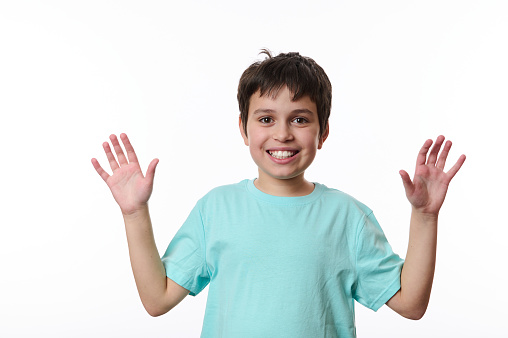 Adorable Caucasian preteen boy, smart school child, wearing a blue t-shirt, shows his hands palms to camera and smiles with a beautiful toothy smile, isolated on white background. Copy space for text