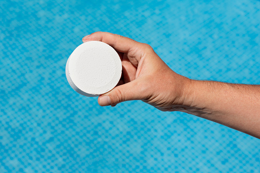 Round white pool chlorine tablet to prevent the appearance of fungi and bacteria. Hand holding pool chlorine tablet over blue water. Large chlorine tablet for floating dispenser