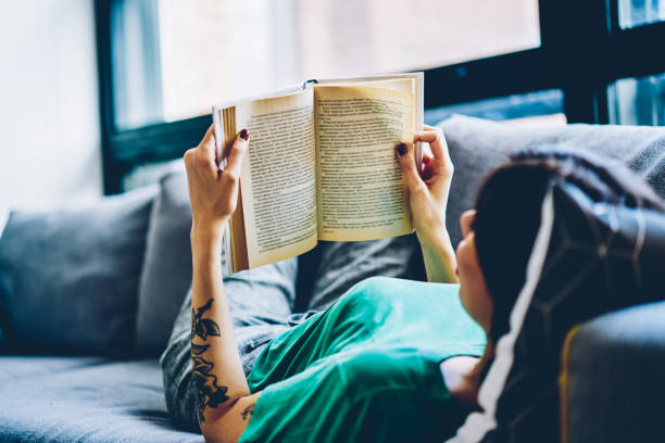 istock Back view of female holding open book on pages resting on comfortable sofa during weekend at home interior, 30s caucasian woman lying on couch reading interesting literature in living room on leisure 1444722130