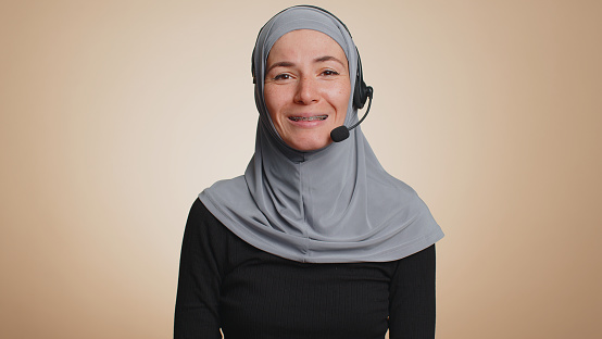 Muslim woman in hijab wearing headset, freelance worker, call center or support service operator helpline, having talk with client or colleague communication support. Arabian girl on beige background