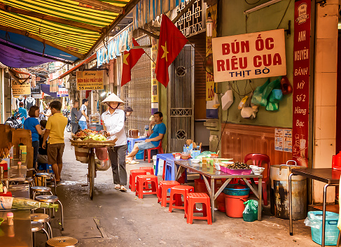 Street vendor passing through Hanoi food market. The narrow street is lined with low chairs and tables