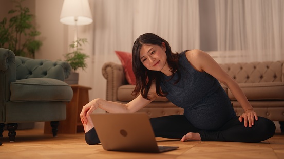 A pregnant woman is sitting on the floor and stretching her body while watching online tutorials on a laptop in the living room at home.