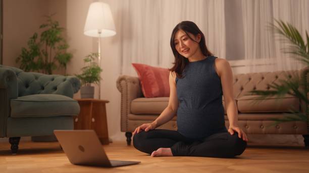 schwangere frau sitzt auf dem boden und streckt ihren körper, während sie online-tutorials auf dem laptop im wohnzimmer zu hause anschaut - floor women sitting yoga stock-fotos und bilder