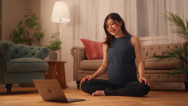 Pregnant woman sitting on floor and stretching her body while watching online tutorials on laptop in living room at home