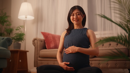 A portrait of a pregnant woman in sports clothing looking and smiling for the camera in the living room at home.