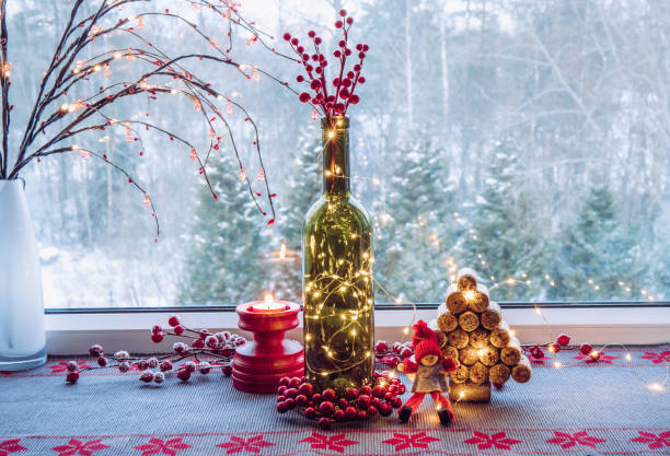 Christmas decoration set with wine bottle filled with micro led party lights and spruce tree made with used wine corks, cute vintage elf figurine, behind is window with snowy countryside  forest.