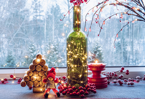 Christmas decoration set with wine bottle filled with micro led party lights and spruce tree made with used wine corks, cute vintage elf figurine, behind is window with snowy countryside  forest.