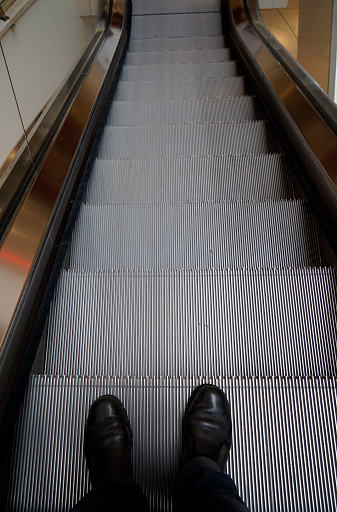 Feet of a man on an escalator, that goes down. Nobody else is there. Photo is made in a large shopping hall in Leverkusen, Germany