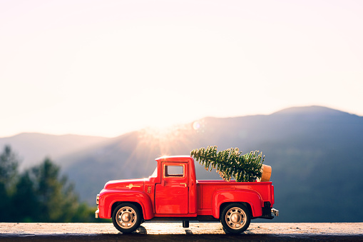 Red retro small car carrying christmas tree in the mountain in the sunbeams of sunrise. Winter holiday concept