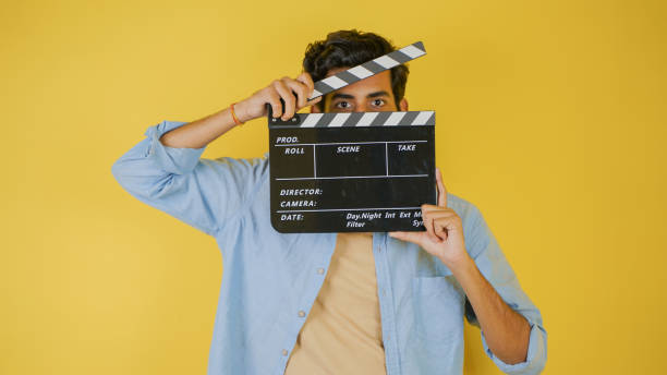 istock Young Asian Indian man standing holding clapperboard shocked, hiding behind clapper board used in film making, isolated on color background studio portrait 1444771453