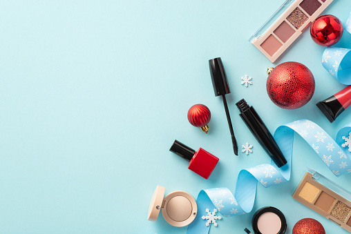 Christmas shopping concept. Top view photo of decorative cosmetics lip gloss nail polish mascara eyeshadow palettes red baubles curly ribbon snowflakes on isolated light blue background with copyspace
