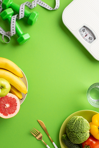 Proper nutrition concept. Top view vertical photo of pates with fruits and vegetables cutlery glass of water scales dumbbells and tape measure on isolated green background with copyspace