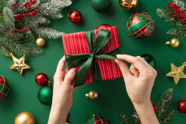 Christmas concept. First person top view photo of woman's hands untying ribbon bow on red giftbox over gold green baubles star ornaments mistletoe pine branches in snow on isolated green background Christmas concept. First person top view photo of woman's hands untying ribbon bow on red giftbox over gold green baubles star ornaments mistletoe pine branches in snow on isolated green background unwrapping stock pictures, royalty-free photos & images