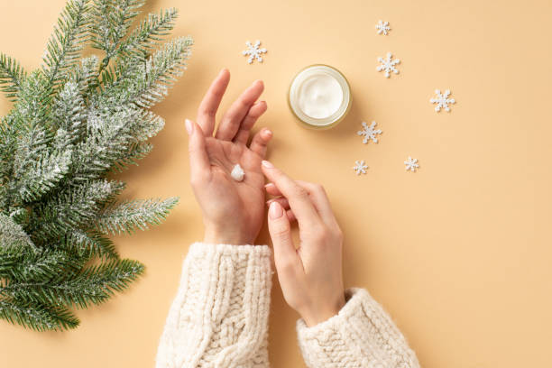winter skin care concept. first person top view photo of girl's hands in white sweater applying cream on her hands from jar fir branches in frost and snowflakes on isolated pastel beige background - christmas tree snow fir tree isolated imagens e fotografias de stock