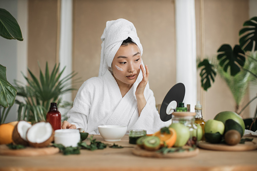 Healthy asian woman in white bathrobe and towel sitting at wooden table with various ingredients preparing natural cosmetics at home mixing mass in bowl while preparing cream and applying on face.