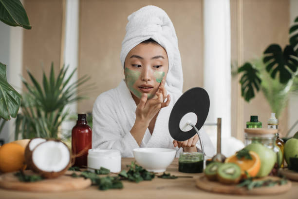 atractiva mujer asiática mirando al espejo sentada en la mesa con ingredientes para cosméticos caseros - facial cleanser fotografías e imágenes de stock