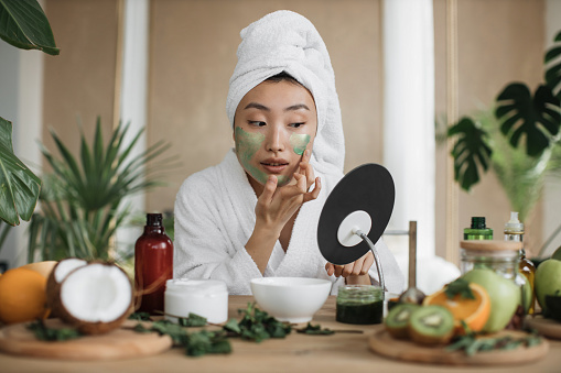 Attractive asian woman looking at mirror sitting at table with ingredients for homemade cosmetics applying green moisturizing mask on her face. Young lady doing anti aging procedures.