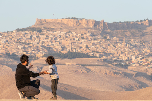 Father showing the historical Mardin landscape to his son. Historical Mardin city center located on the top of the mountain. 
Father and son looking at the city view of Mardin consisting of stone houses. Shot with a full frame camera.