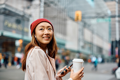 Happy Asian woman using smart phone while carrying takeaway coffee and walking on the street.