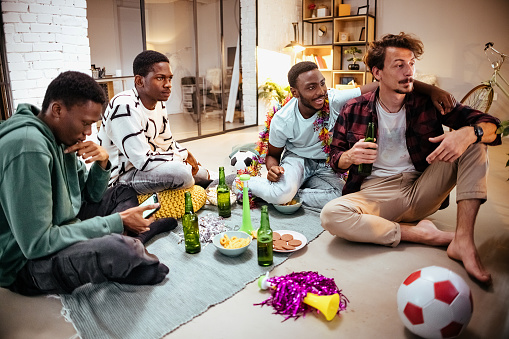 Male friends sit on the living room floor and watch a football game together