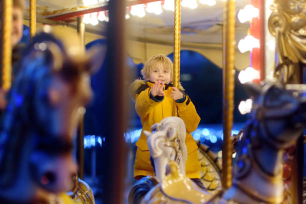 Cute blonde boy enjoying Christmas fair. Little child riding on a vintage carousel (merry go round). Outdoors entertainment activity for children on winter holidays stock photo