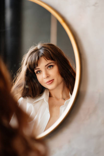 красивая женщина в белой рубашке смотрит на свое отражение в круглом зеркале. - mirror women human face sensuality стоковые фото и изображения